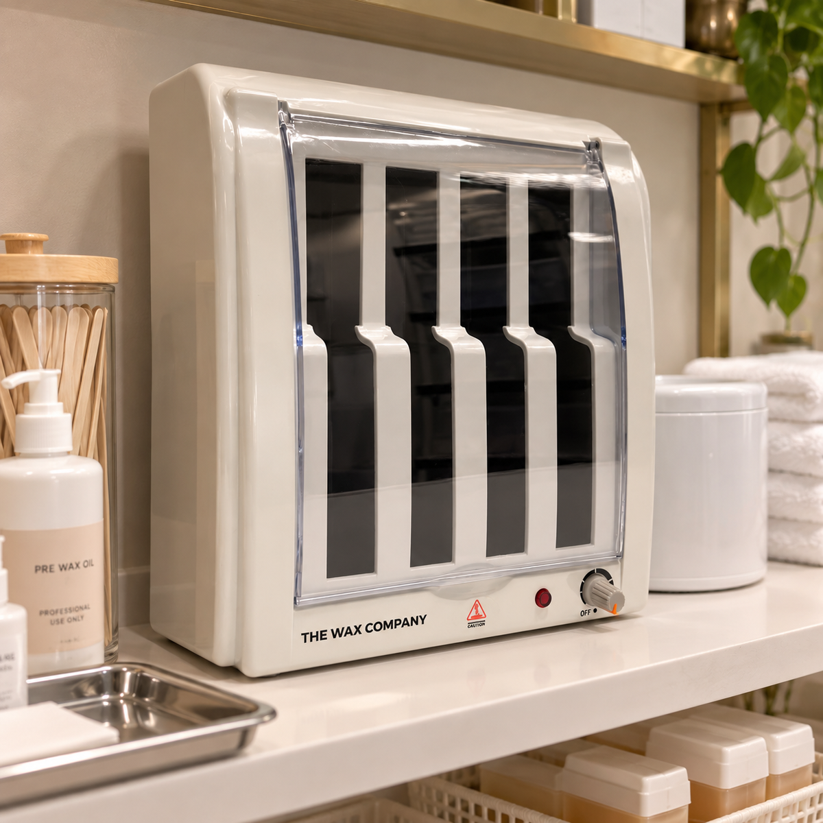 4 bay roller wax warmer by The Wax Company sits on a bathroom or spa counter, surrounded by skincare products, folded white towels, and a gold-framed shelf.